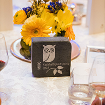 Nahaufnahme einer Preistafel mit Eulenmotiv, umgeben von gelben Blumen, Weingläsern und einer sanft beleuchteten Tischdekoration.