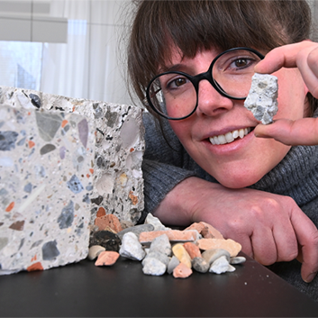 Eine Person lächelt, während sie ein kleines Trümmerstück neben einem größeren Block und weiteren Fragmenten auf einem Tisch hält und zeigt.