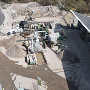 Luftaufnahme eines Industriegeländes mit Kieshaufen, Maschinen und einer großen überdachten Fläche, umgeben von Bäumen.
