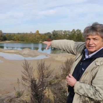 Eine Person deutet auf einen See mit umliegenden Bäumen und sandigem Gelände unter einem teilweise bewölkten Himmel.