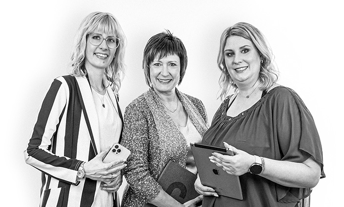 Drei lächelnde Frauen mit technischen Geräten in der Hand stehen vor einem schlichten Hintergrund auf einem Schwarzweißfoto.