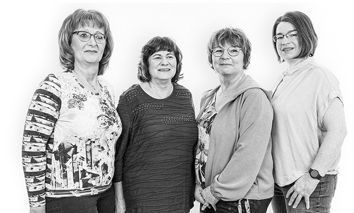 Vier Frauen stehen in einer Reihe und lächeln in die Kamera, auf einem Schwarzweißfoto.