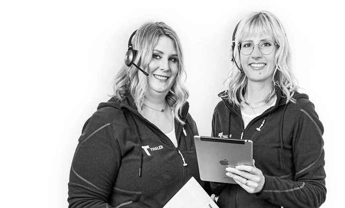 Zwei Frauen mit Headsets und Thule-Jacken, lächeln und halten vor einem schlichten Hintergrund ein Tablet und Papiere in der Hand.