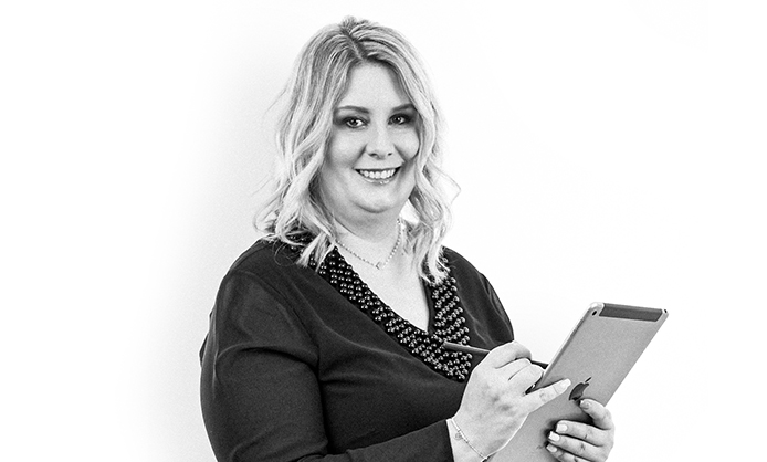 Eine Frau mit mittellangem Haar lächelt, während sie vor einem schlichten Hintergrund ein Tablet und einen Stift hält.