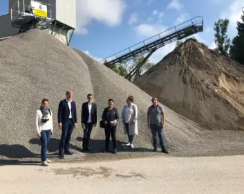Sechs Menschen stehen unter sonnigem Himmel vor großen Kies- und Sandhaufen.